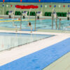 The Swimming Pool Mat lines the pool edge, providing a blue non-slip surface beside clear water, stainless steel handrails, swimmers in the background, green chairs nearby, and colourful triangular flags hanging above.