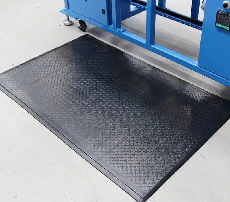 A black anti-fatigue mat with a textured chequer plate surface is placed on a light grey floor next to blue industrial equipment.